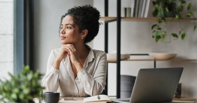Thoughtful business professional seated at a desk in a modern office, gazing out a window in a reflective pose with a laptop nearby.
