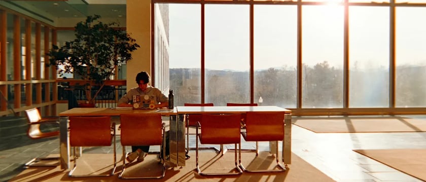 A person works on a laptop at a table beside large sunlit windows, with empty chairs nearby.