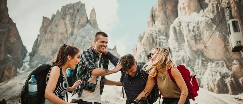 Four people with backpacks laughing together beneath rocky mountain peaks.
