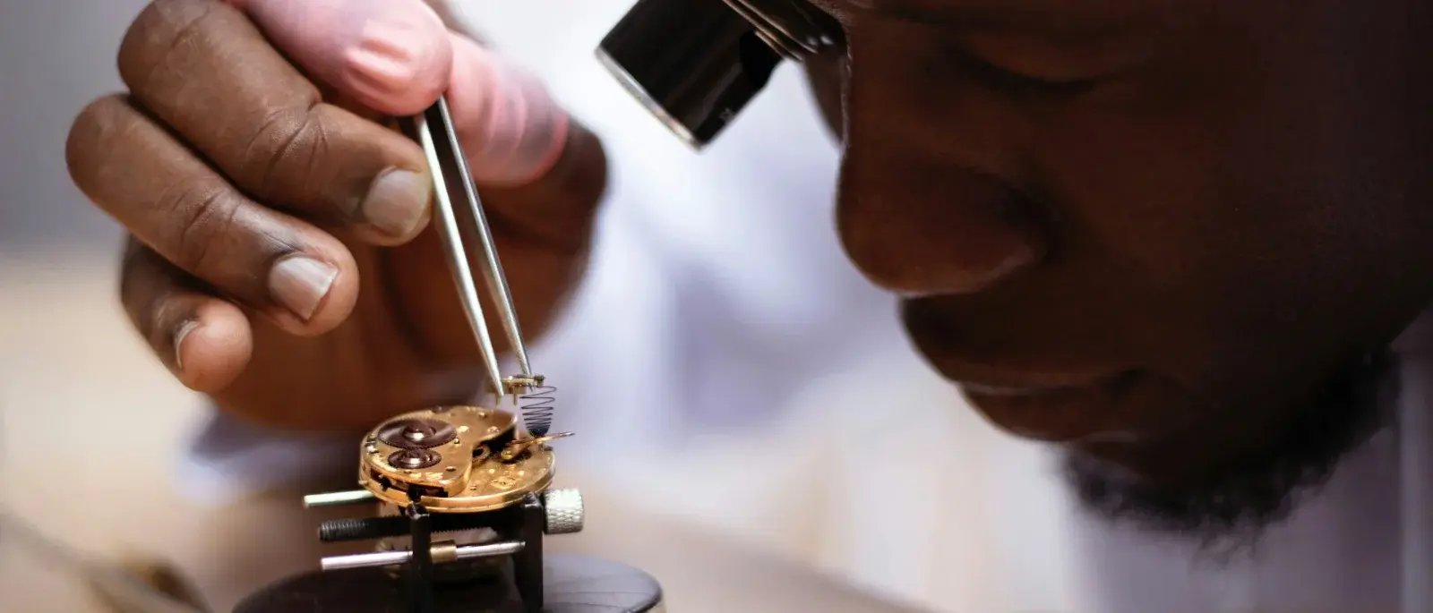 A watchmaker uses an eyepiece and tweezers to work on a small watch mechanism.