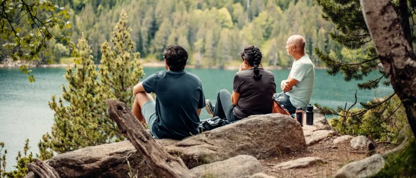 three people sitting on rocks beside a forest-lined lake