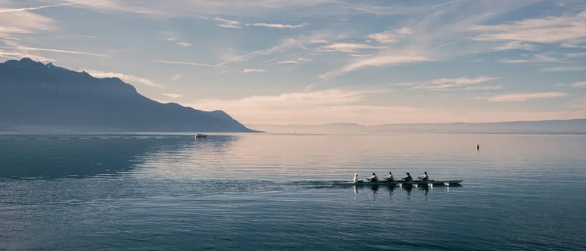 A rowing crew crossing a calm lake with mountains in the background