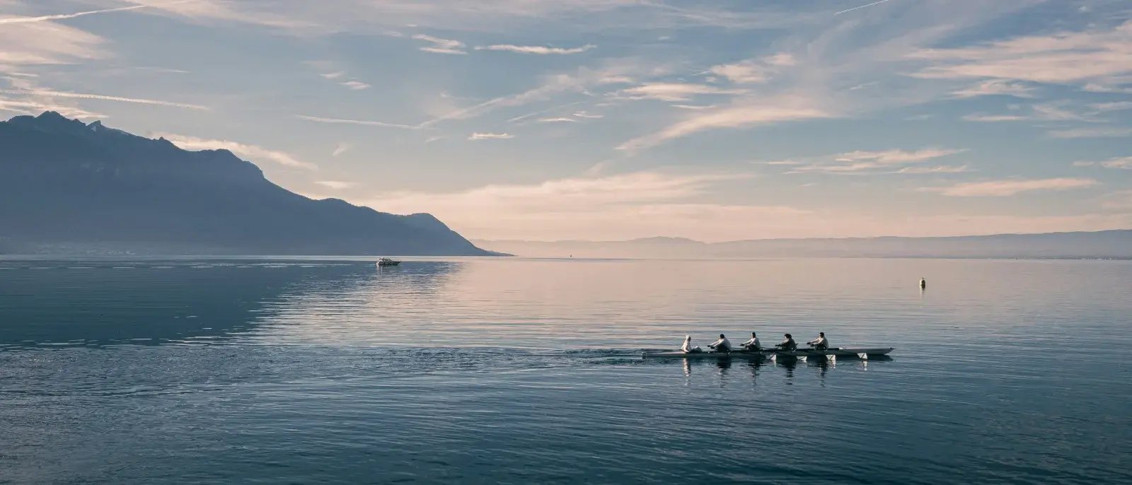 A rowing crew crossing a calm lake with mountains in the background