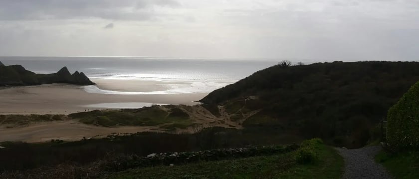 A coastal path leads towards a sandy beach, with sunlight breaking through clouds over the sea.