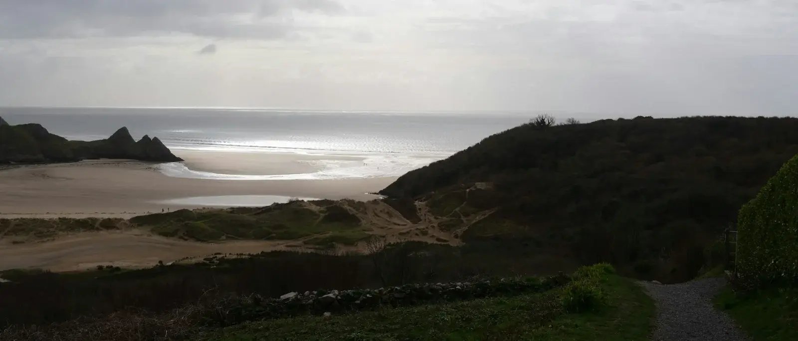 A coastal path leads towards a sandy beach, with sunlight breaking through clouds over the sea.
