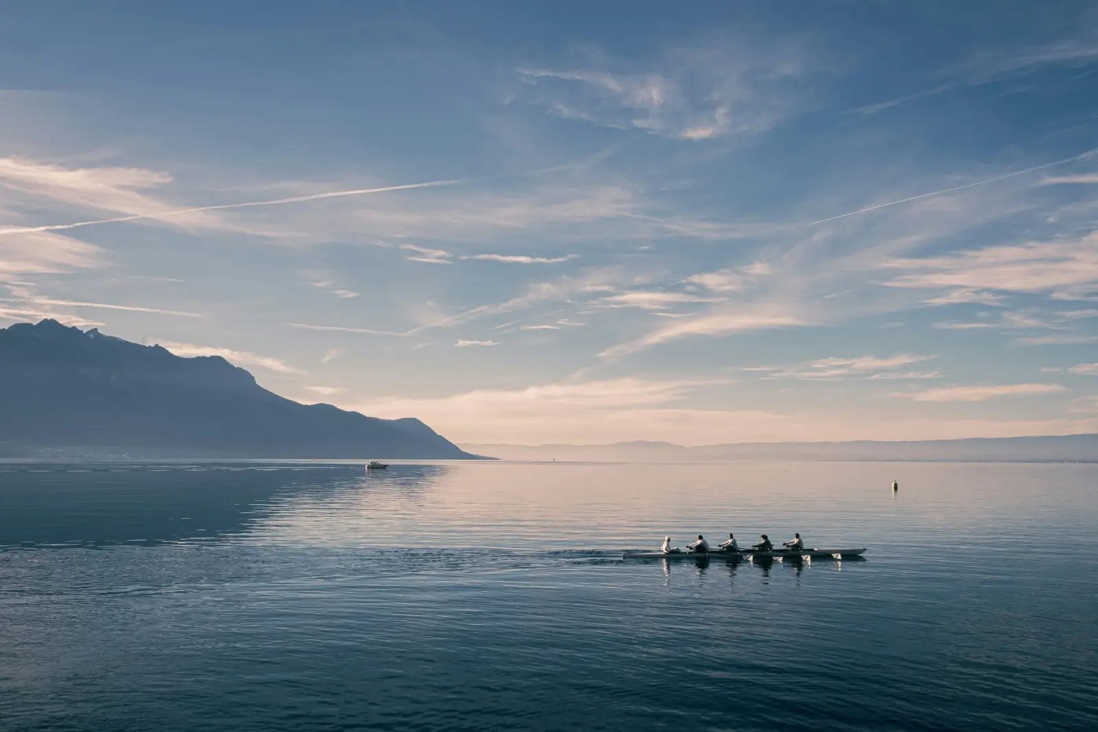 A rowing crew moves together across a calm lake beneath a mountain skyline.