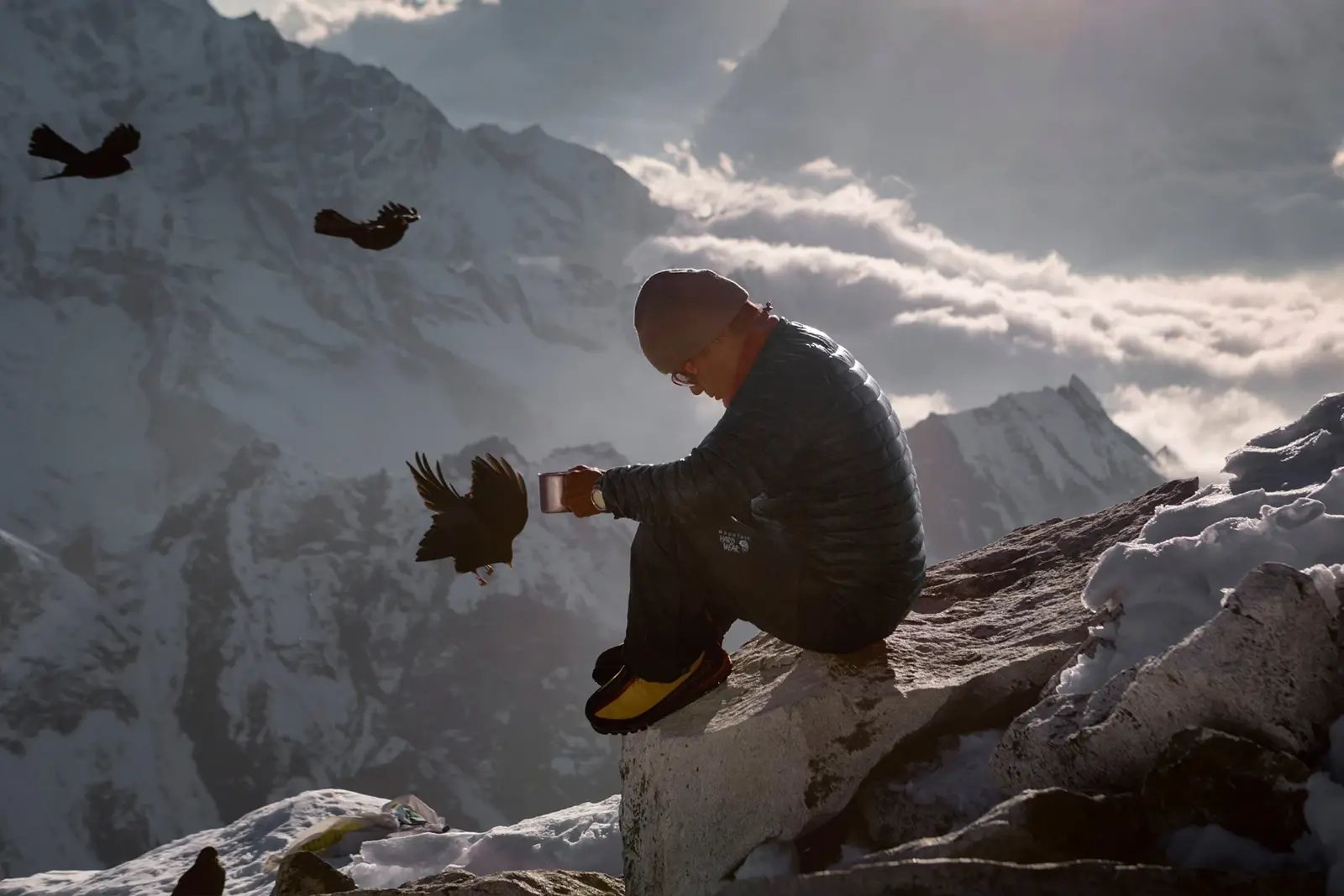 A mountaineer seated on a snowy ridge holds out a cup towards three birds, with snow-covered mountains behind.